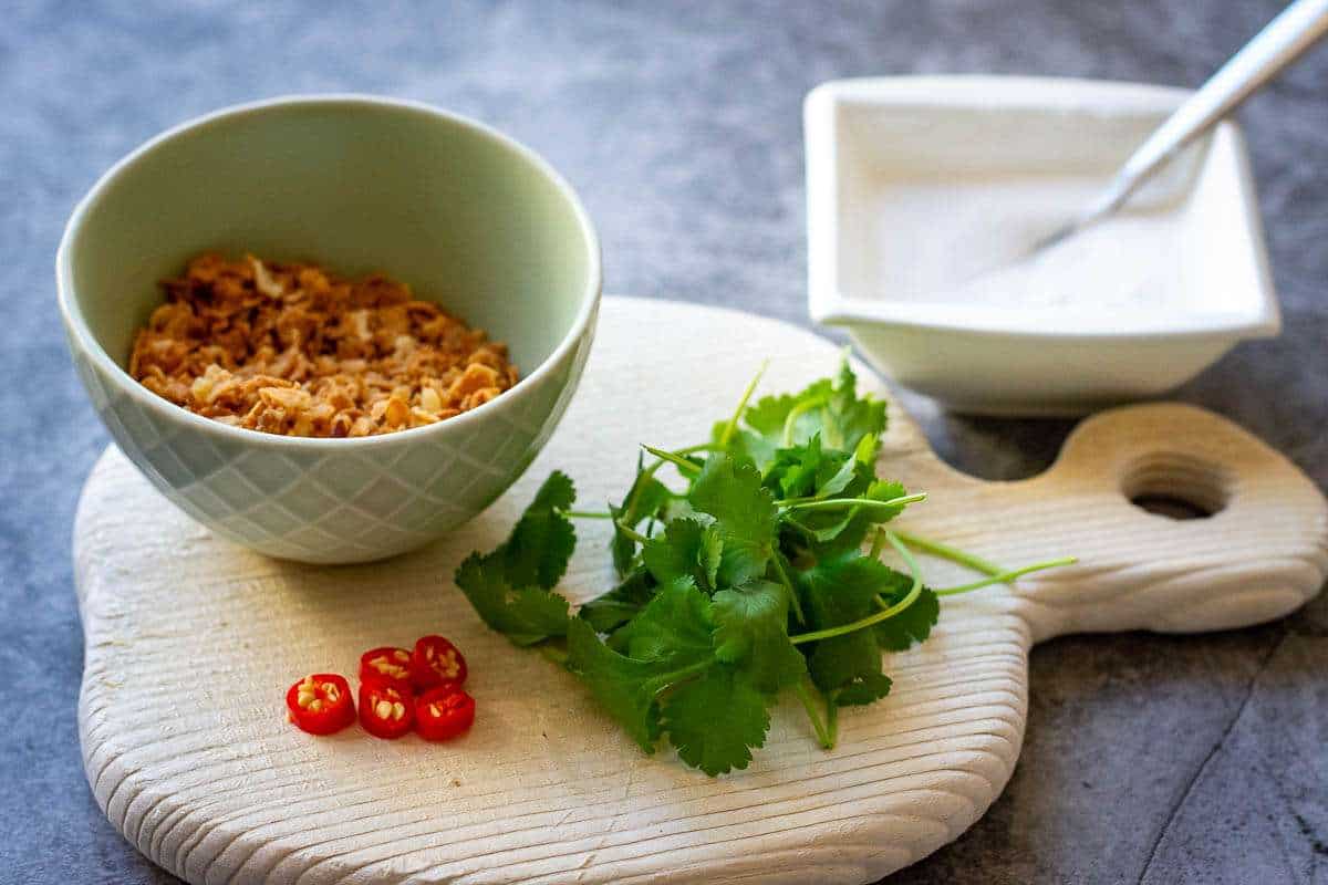 Coriander leaves, sliced red chilli pepper, a small bowl of crispy onions and a square white bowl of reserved coconut milk ready for garnishing the soup.