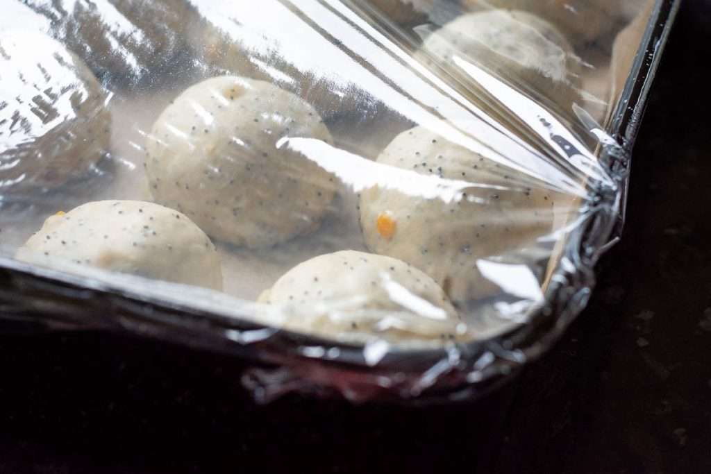 Baking tray with high rims with hot cross buns in it covered with cling film.