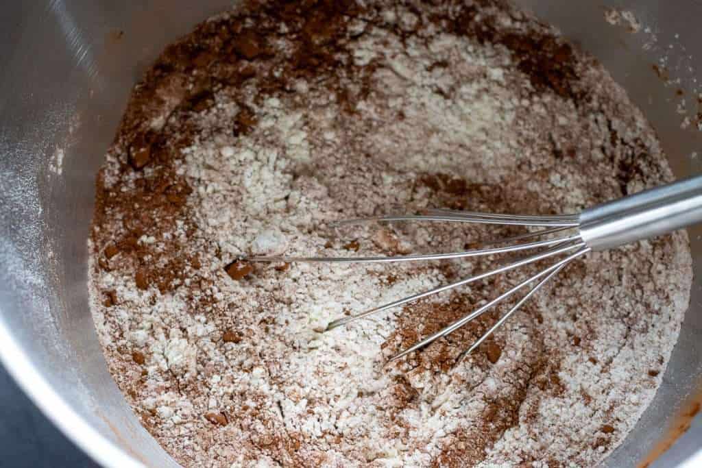 Mixing dry ingredients for sourdough chocolate hot cross buns in a bowl of a stand mixer using a balloon whisk.