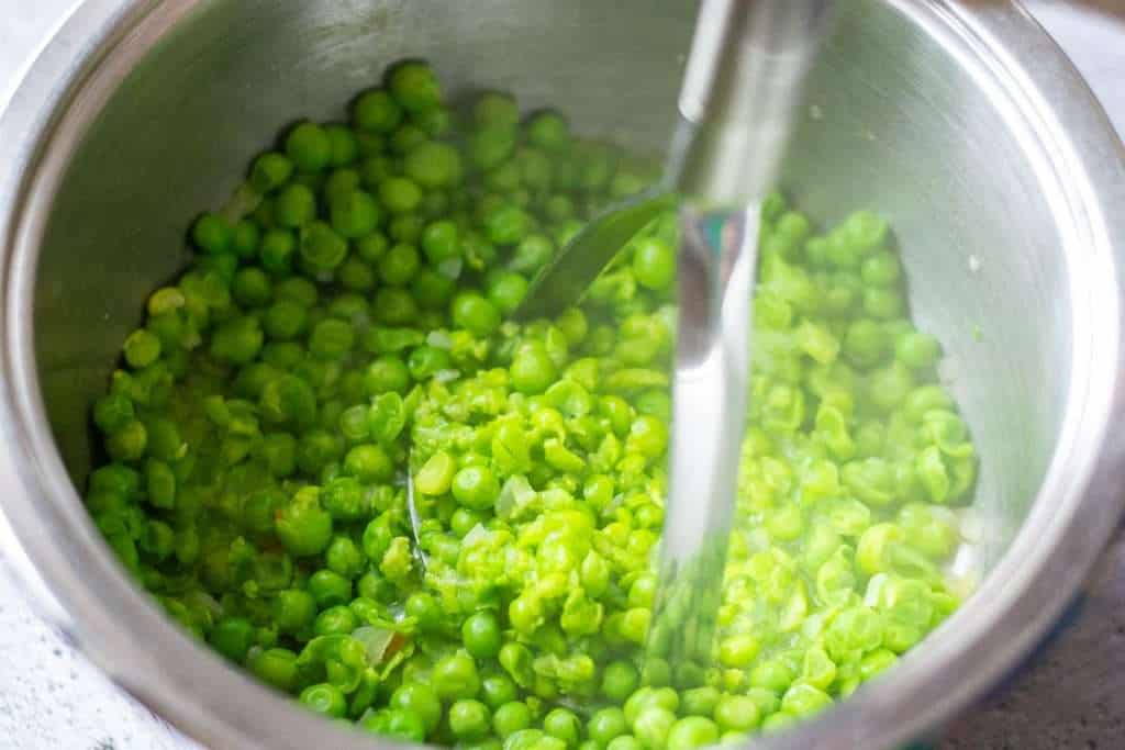 Mashing cooked green peas with a potato masher.