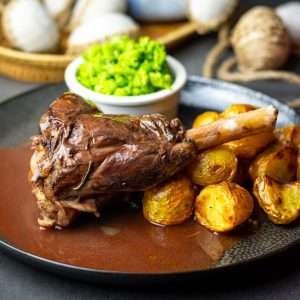 Slow cooker lamb shank in a pool of red wine sauce on a black plate with roast potatoes and minted peas.