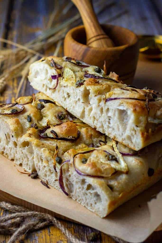 Sliced and stacked sourdough focaccia with fennel and capers on a wooden board.