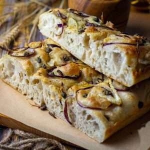 Sliced and stacked sourdough focaccia with fennel and capers on a wooden board.