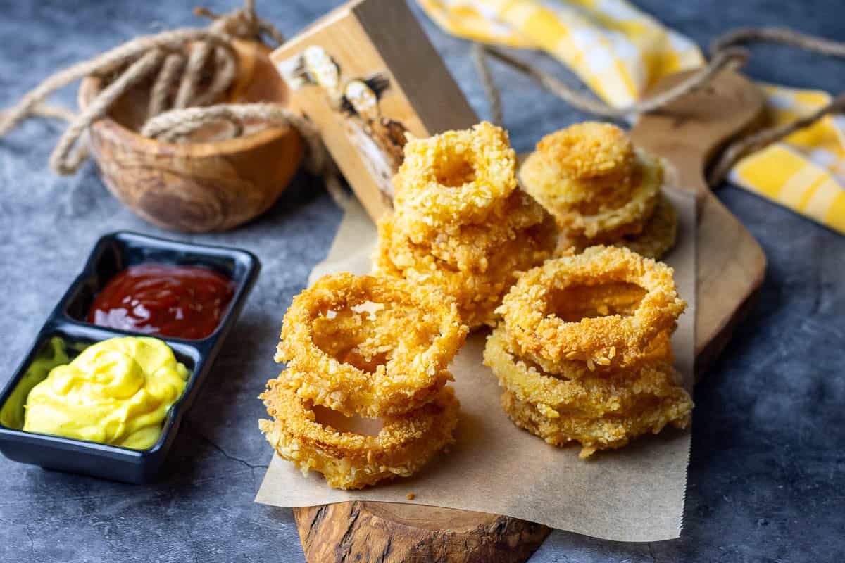 three stacks of sourdough onion rings on a parchment paper lined wooden board next to two dips.