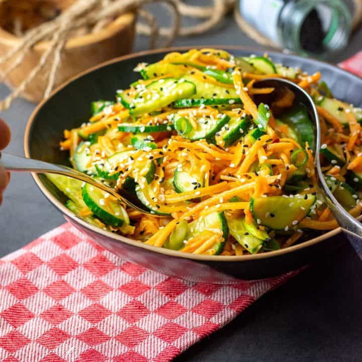 Carrot and cucumber salad with sesame seeds on top in a black bowl.