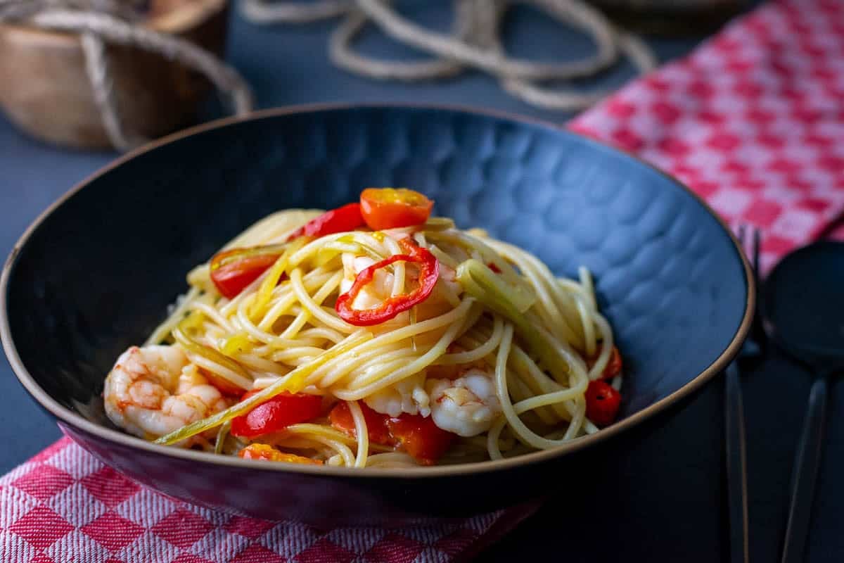 A black bowlful of fennel, prawn and cherry tomato pasta on a table with red teatowel on it.