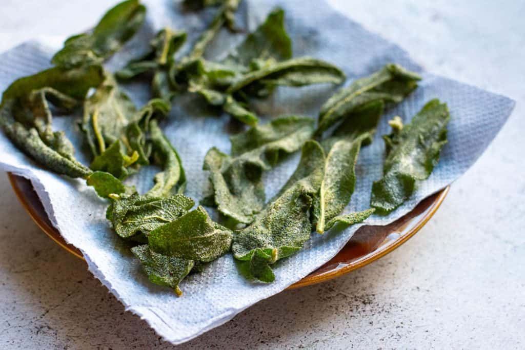 Crispy sage leaves arranged on a paper towel lined plate.