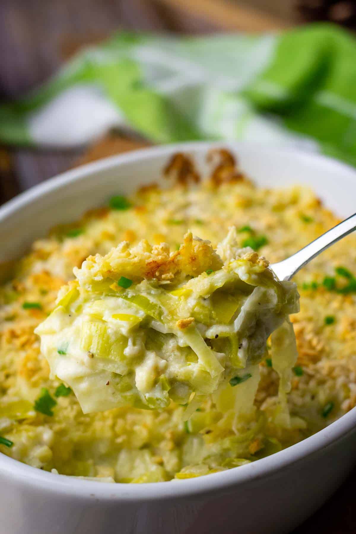 A spoonful of creamy cheesy leeks being lifted out of a casserole dish.