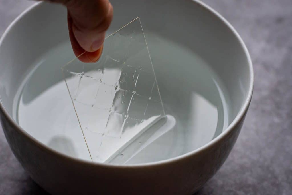 Adding a sheet of leaf gelatine to cold water in a white bowl.