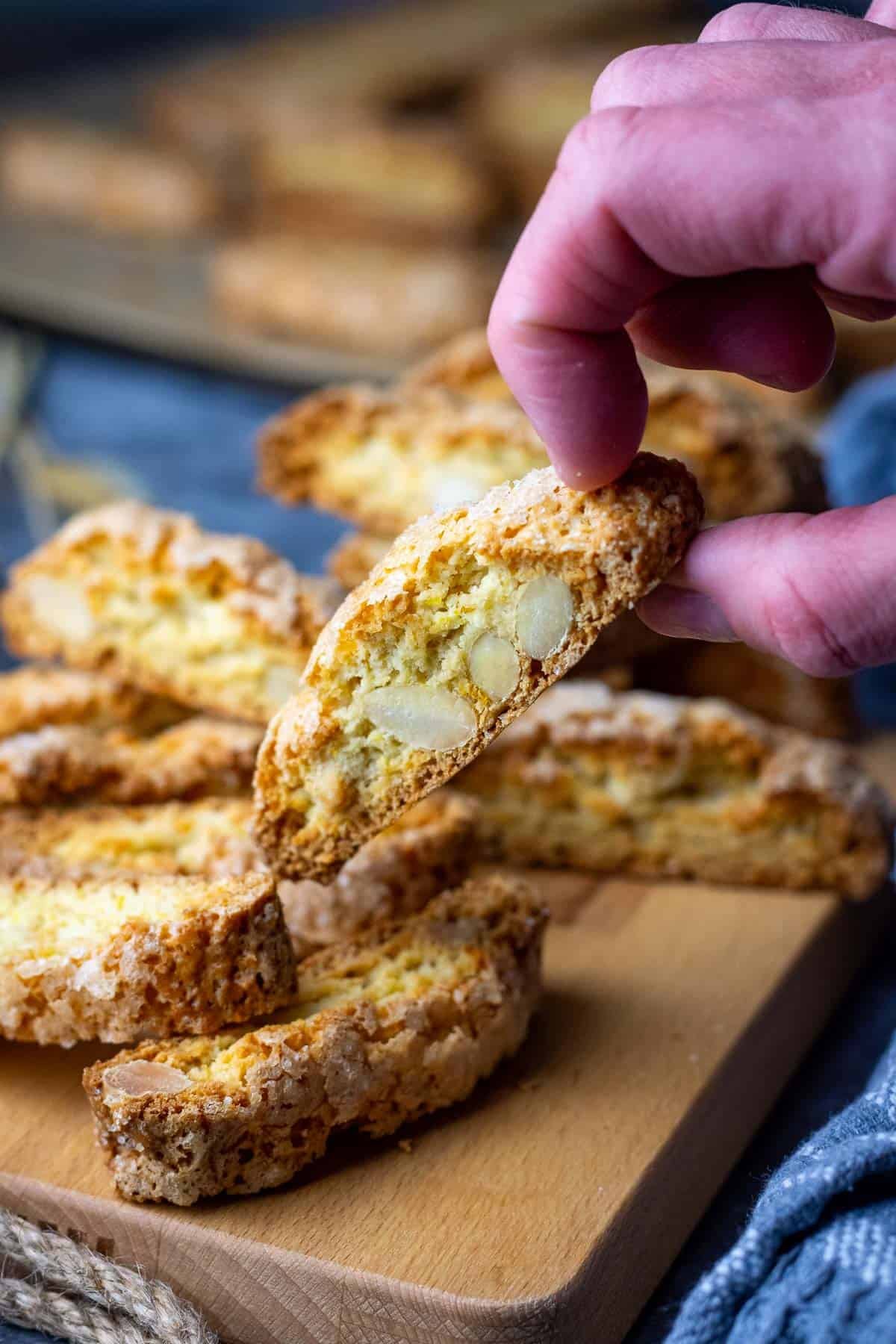 Almond sourdough biscotti on a wooden board with one being picked up by a hand.