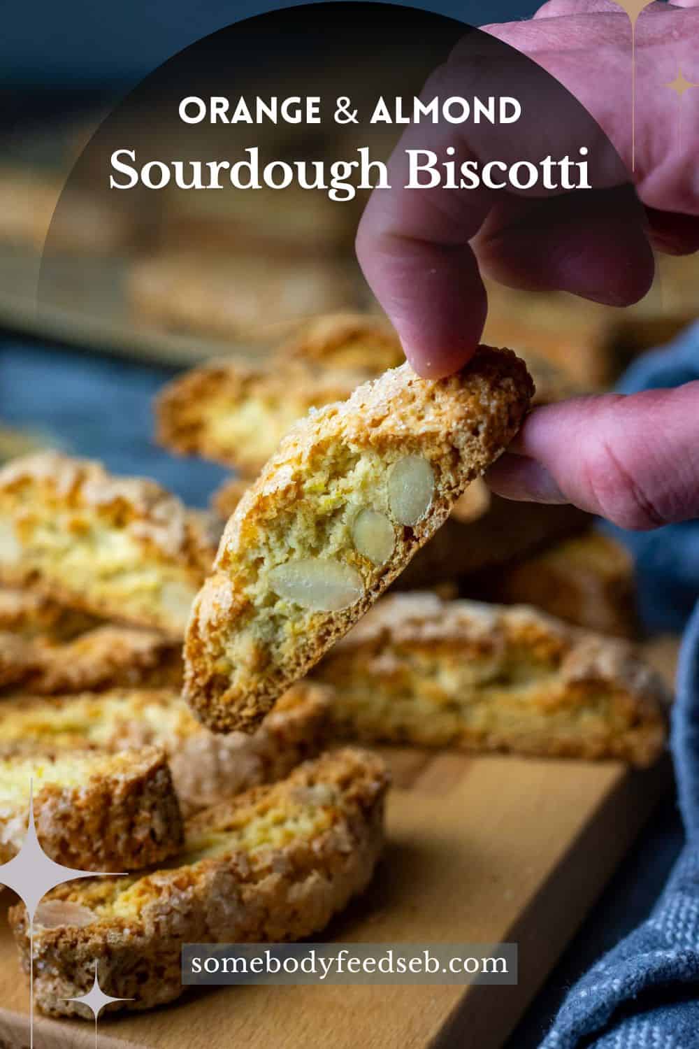 orange and Almond sourdough biscotti on a wooden board with one being picked up by a hand. Pin for Pinterest.