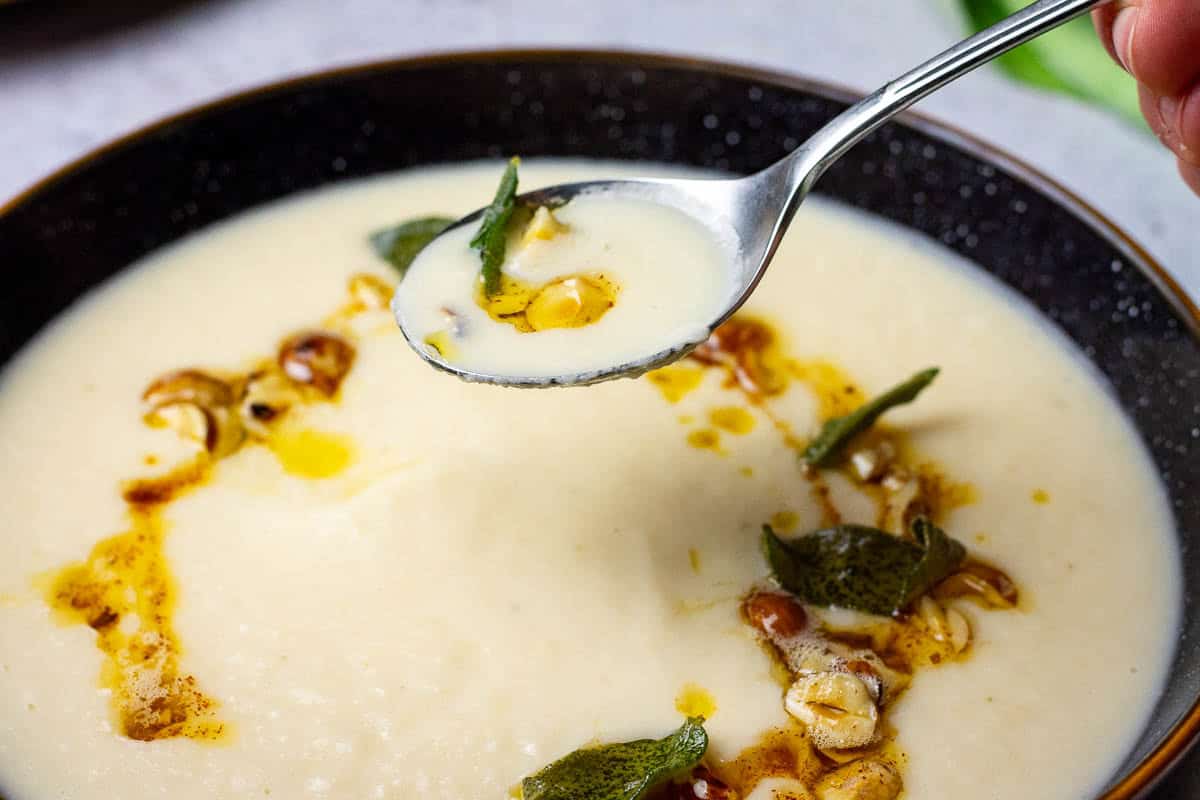 A spoonful of creamy celeriac soup being picked up form a bowlful with a swirl of browned butter, hazelnuts and crispy sage.