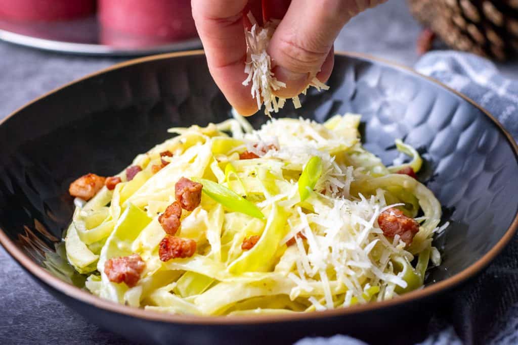 Cabbage alfredo with pancetta in a black bowl being topped with more parmesan.