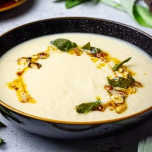 Creamy Celeriac Soup with browned butter, hazelnuts and crispy sage on top in a black bowl.