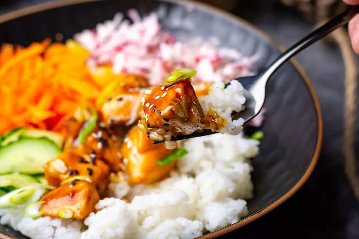 A piece of teriyaki salmon being icked up from a poke bowl.