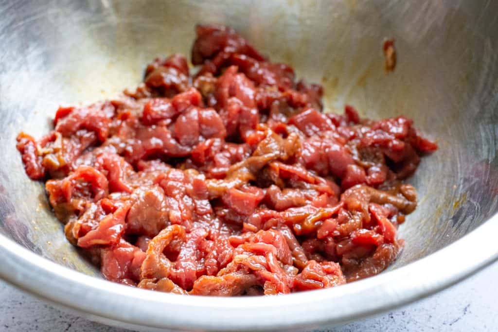 Beef strips in marinade in a large metal bowl.