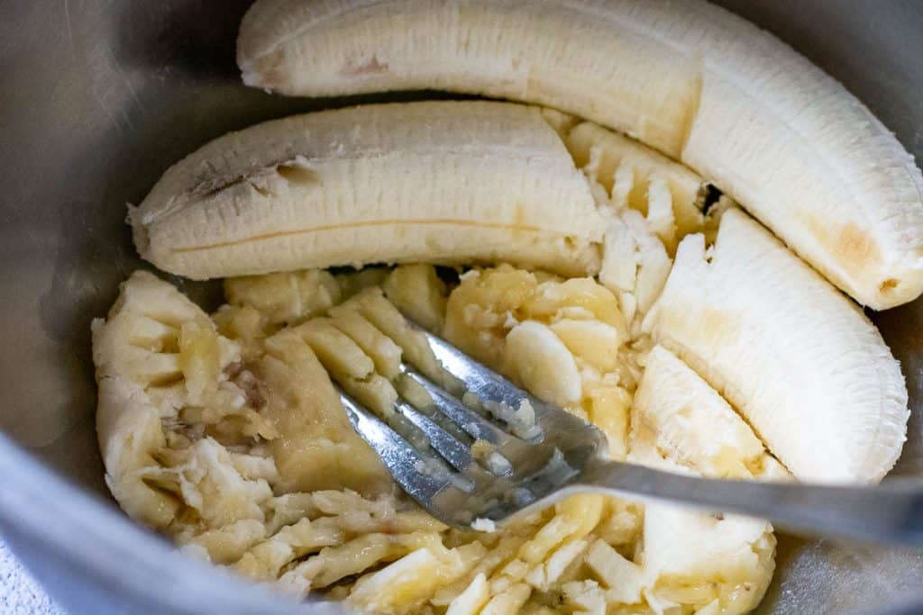Mashing bananas with a fork in a metal bowl.