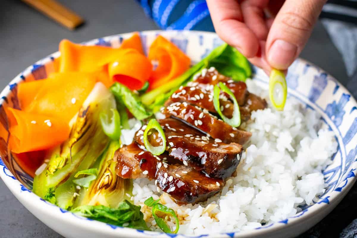 Teriyaki pork tenderloin sliced on a bed of rice and drizzled with extra teriyaki sauce, being garnished with sliced spring onions.