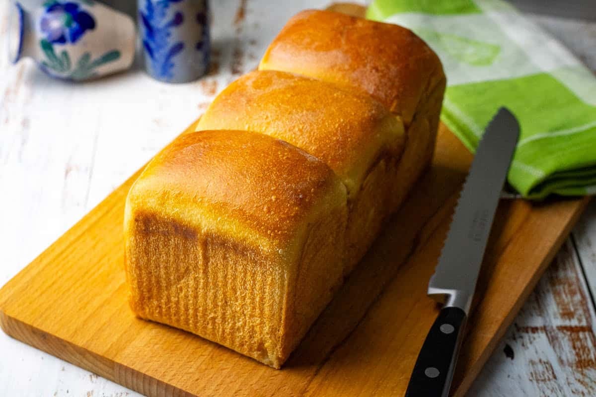 Golden brown evaporated milk sourdoughsandwich bread on a wooden boar with a bread knife next to it.