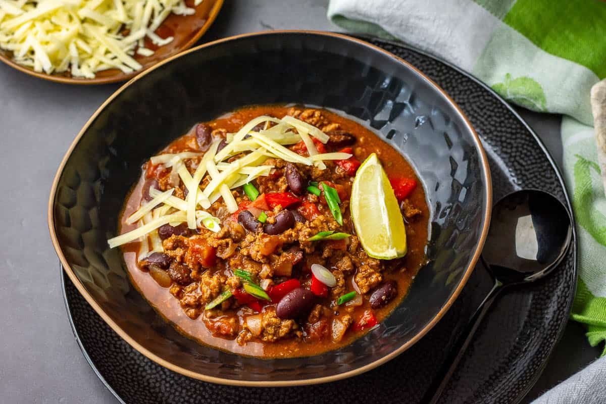 A bowlful of slow cooked chilli con carne topped with cheese, spring onions and a lime wedge.