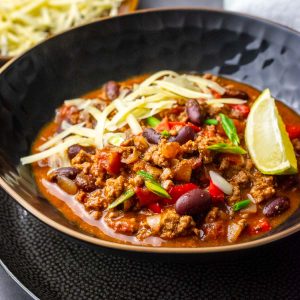 A bowlful of slow cooked chilli con carne topped with cheese, spring onions and a lime wedge.