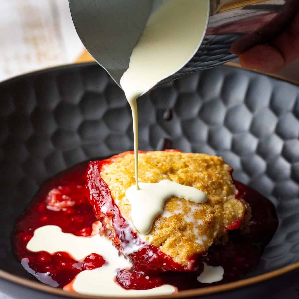 Cream being poured on sourdough strawberry cobbler in a black bowl.