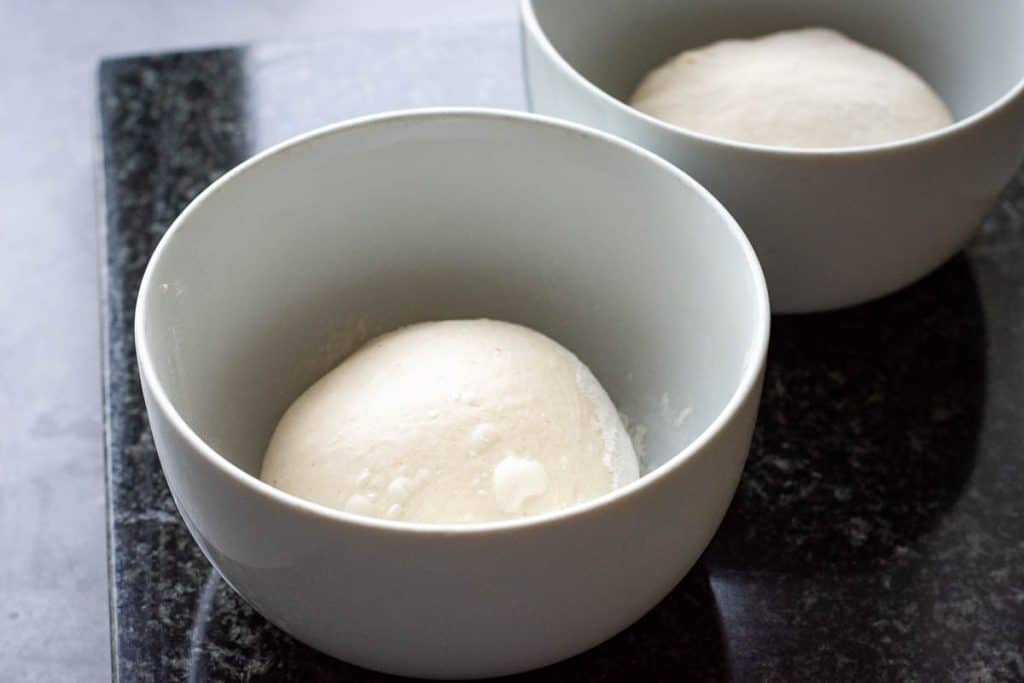 Two floured white bowls with a ball of pizza dough in each.