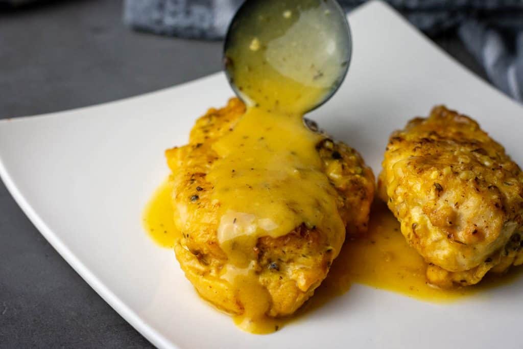 Spooning honey mustard sauce over the chicken breasts on a white plate.