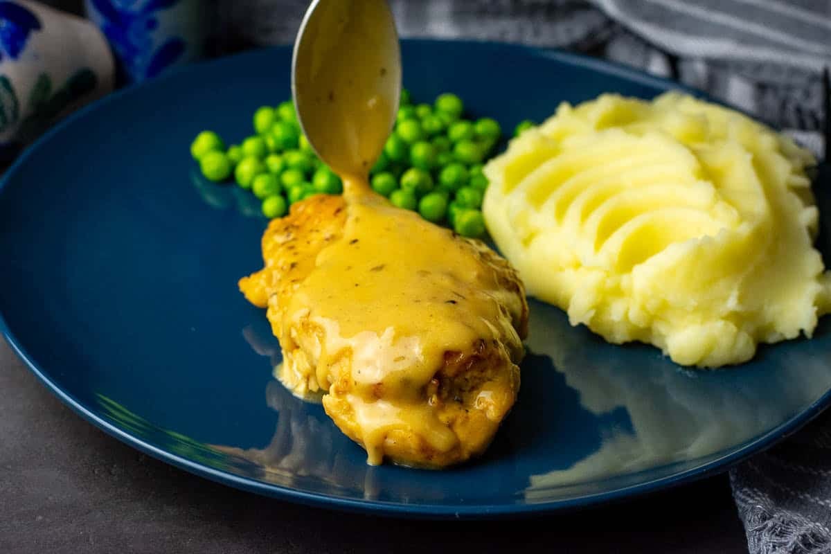 Spooning thickened hoey mustard sauce over the chicken breast served alongside mashed potatoes and green peas.