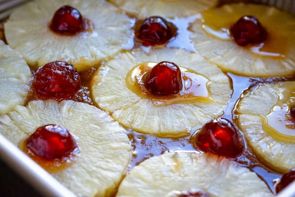 Pineapple slices and glace cherries arranged on top of the butter and sugar on the base of a baking pan.