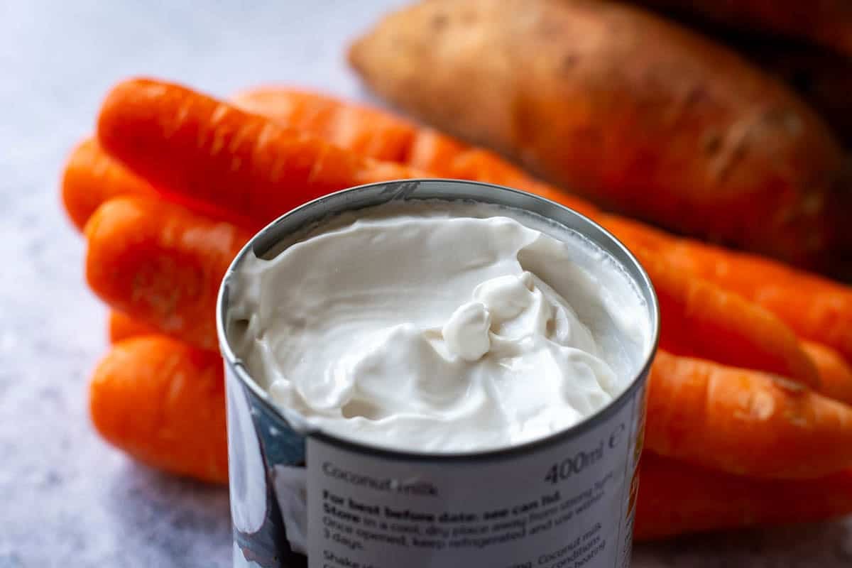 A tin of creamy and rich coconut milk with carrots and sweet potatoes in the background.