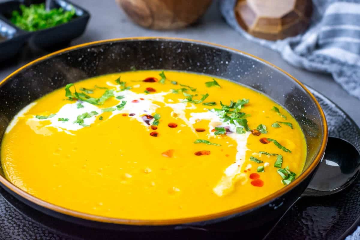 Coconut Milk, Carrot and Sweet Potato Soup with mor ecoconut milk, coriander, crungy chilli oil on top in a black bowl.