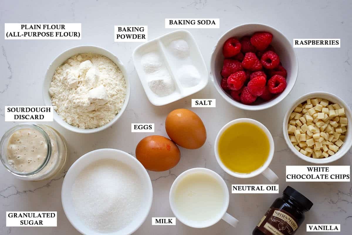 Labelled ingredients for sourdough muffins with raspberries and white chocolate chips on a light quartz kitchen surface.