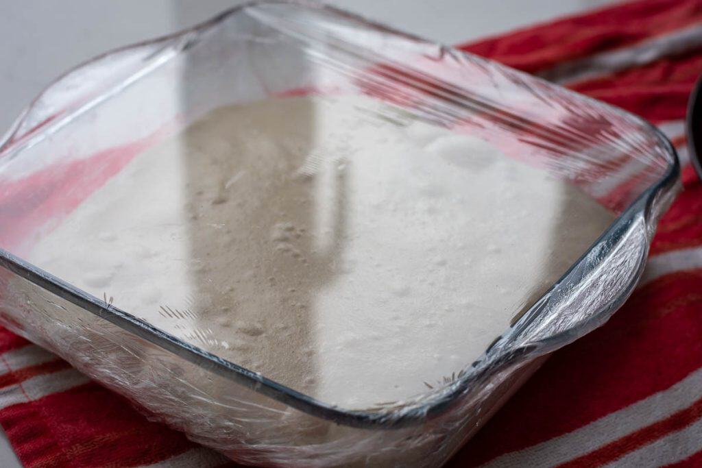 Sourdough focaccia dough in a glass dish covered with plastic wrap amidway through bulk fermentation.