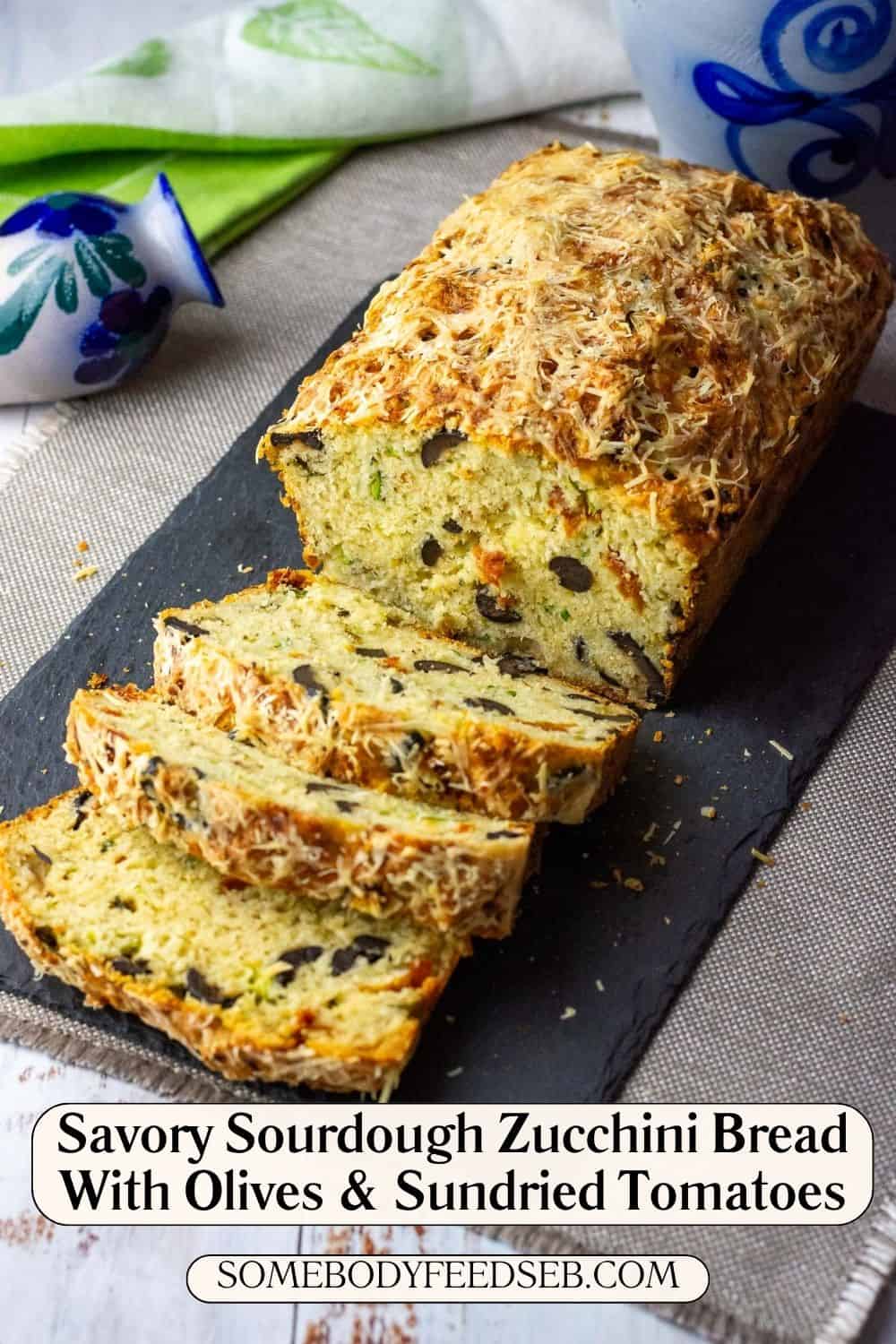 Savoury Mediterranean sourdough zucchini bread loaf with olives and sundried tomatoes sliced on a black slate board. Pin for Pinterest.