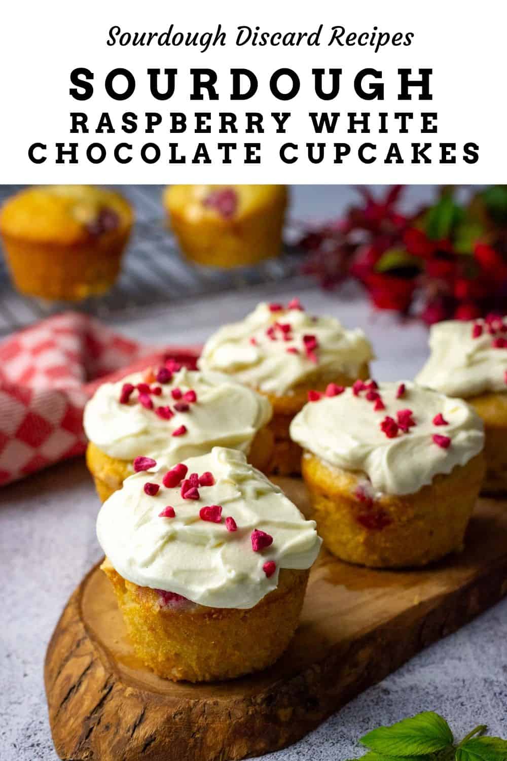 Raspberry and white chocolate sourdough cupcakes with white chocolate cream cheese frosting and freeze dried raspberries on top arranged on a wooden board. Pin for Pinterest.