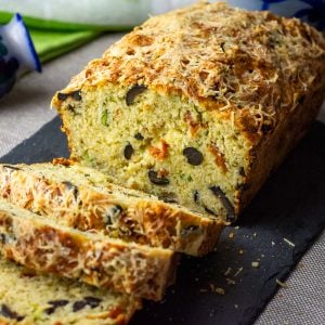 Savoury Mediterranean sourdough zucchini bread loaf with olives and sundried tomatoes sliced on a black slate board.
