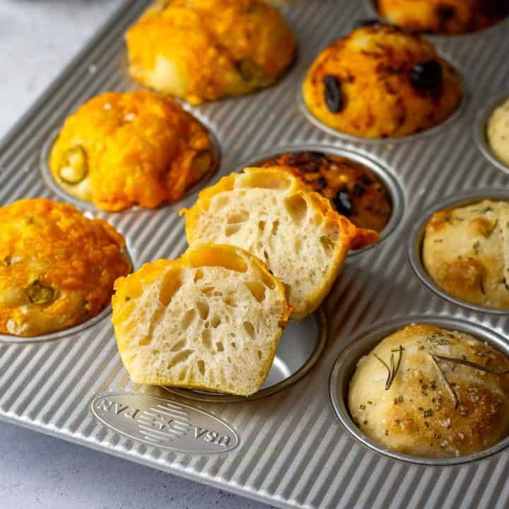 A muffin pan with 12 sourdough focaccia muffins with three different toppings, one muffin is cut in half showing airy interior.
