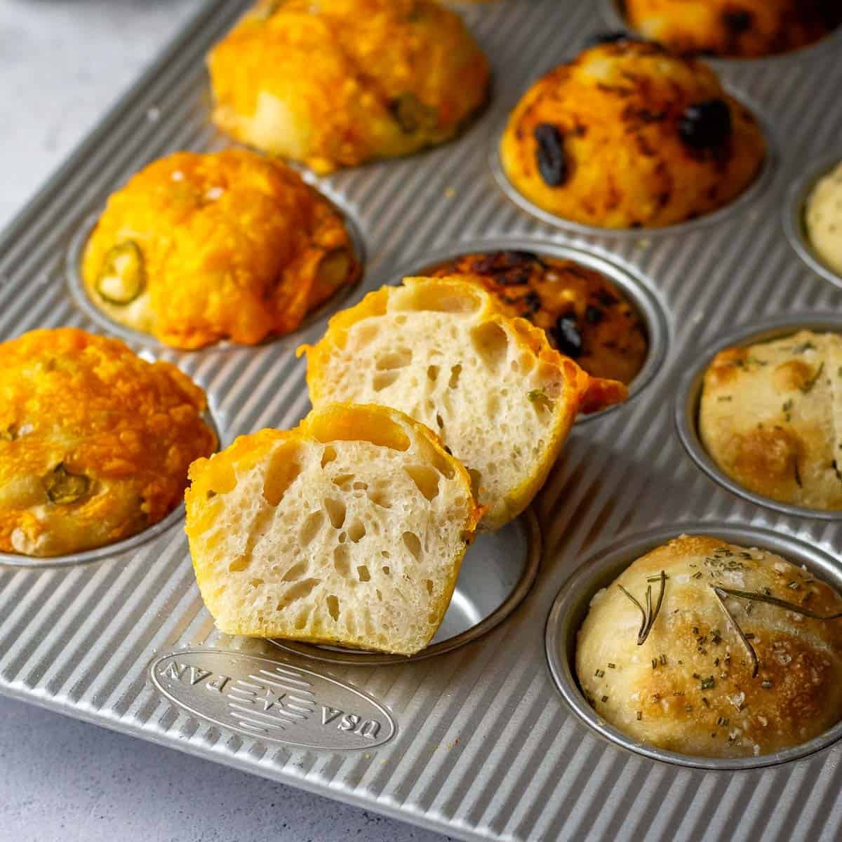 A muffin pan with 12 sourdough focaccia muffins with three different toppings, one muffin is cut in half showing airy interior.