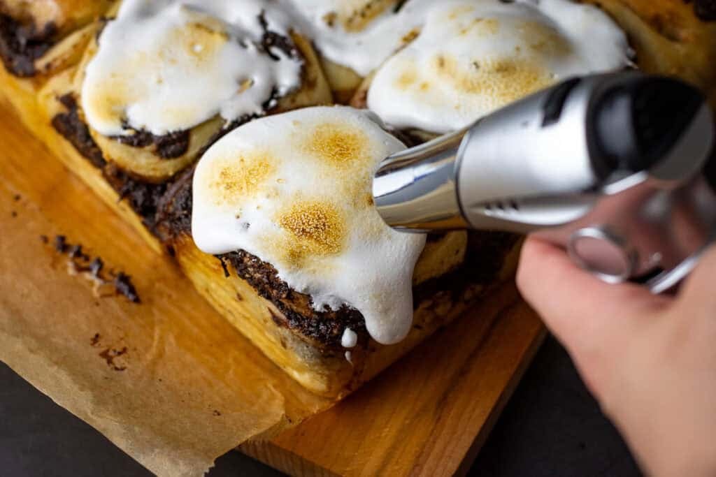 Sourdough s'mores rolls topped with marshmallow fluff being toasted with a kitchen blow torch.