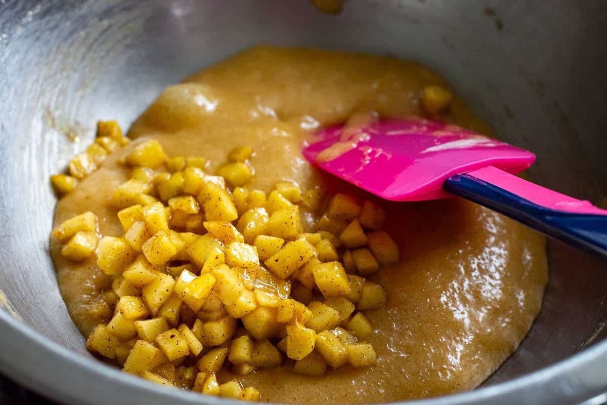 Cubed apples being mixed into wet ingredients with a pink rubber spatula.