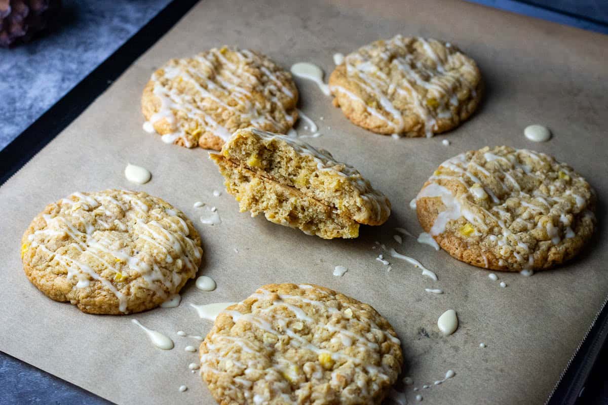 Sourdough discard apple cookies on a baking sheet with one of them broken in half exposing chewy interior studded with apple pieces.