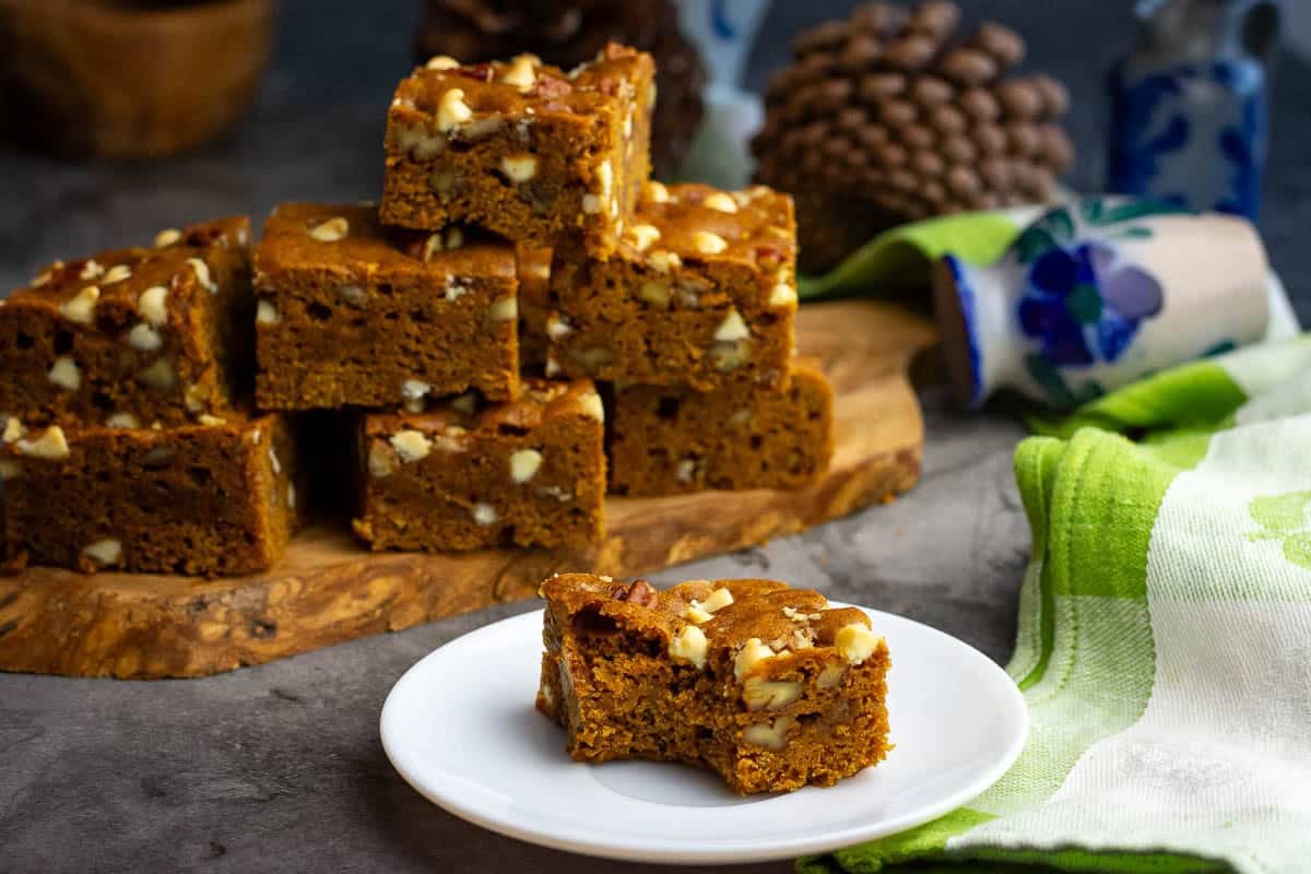 A sourdough pumpkin blondie square bitten into on a small white plate with a stack of sourdough pumpkin blondies in the background.